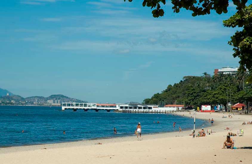 Praia da Bica, na Ilha do Governador, Rio de Janeiro, com faixa de areia clara, mar calmo e banhistas aproveitando o dia ensolarado.
