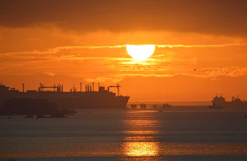 Navios atracados vistos ao pôr do sol na Baía de Guanabara, com o céu alaranjado refletido sobre o mar.