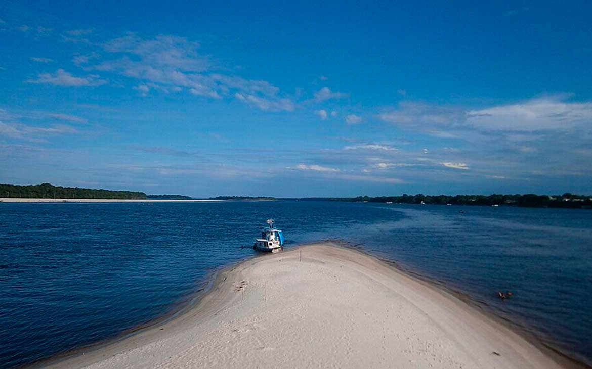 Faixa de areia branca na Ilha do Governador, Rio de Janeiro, avançando sobre o mar com um barco atracado e céu azul ao fundo.