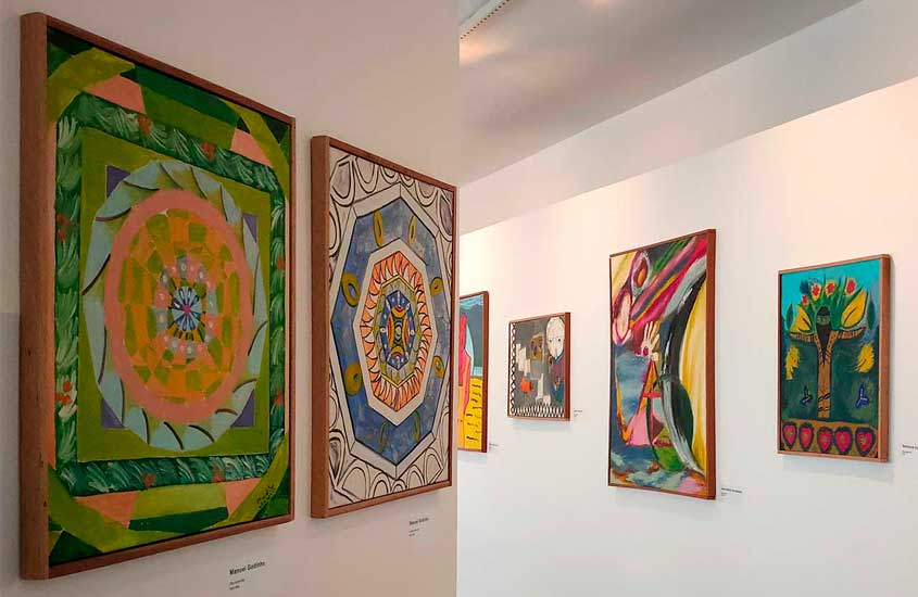 Exposição de arte em museu, com pinturas coloridas em diferentes estilos expostas em paredes brancas iluminadas.