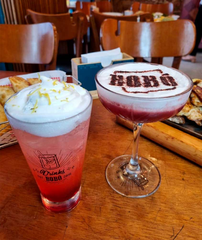 Dois drinks coloridos sobre mesa de madeira em bar, um em copo alto com espuma e raspas de limão e outro em taça decorada com a palavra 'BOBO' no topo de espuma.