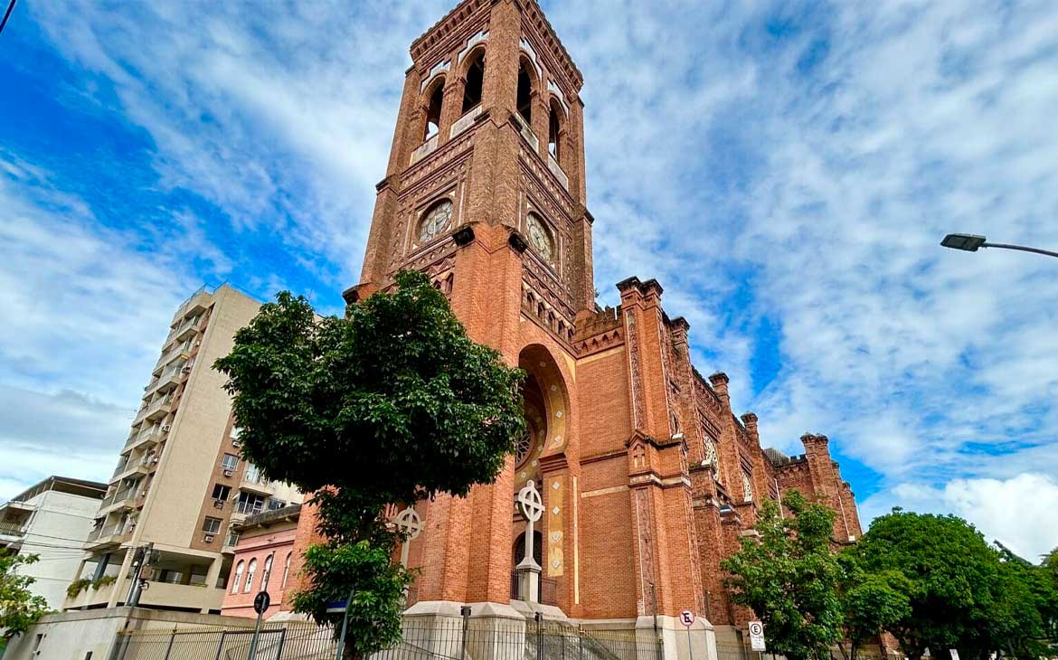 Fachada imponente da Basílica Coração de Maria, em tijolos aparentes, com torre alta e detalhes arquitetônicos históricos, sob céu azul com nuvens.