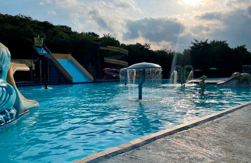 Piscina ampla em parque aquático, com brinquedos infantis, escorregadores e toboáguas ao fundo, iluminada pela luz do sol entre nuvens.