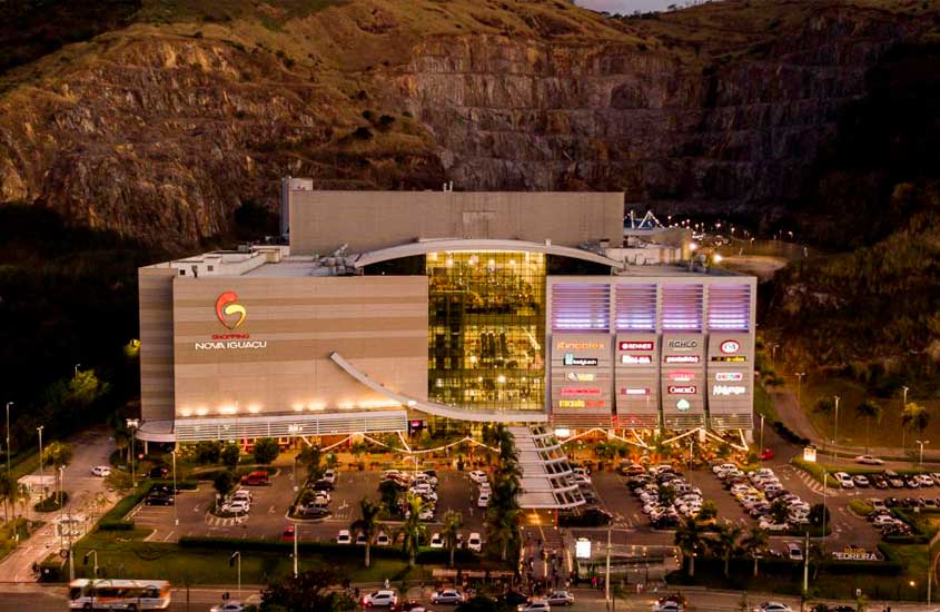 Vista aérea do Shopping Nova Iguaçu iluminado ao anoitecer, com estacionamento cheio e cercado por montanhas de pedra ao fundo.