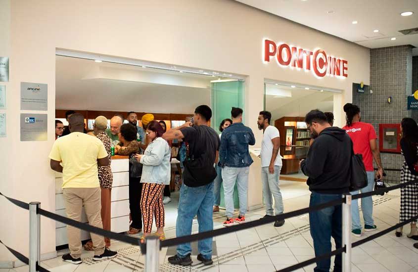 Grupo de pessoas aguardando em fila na entrada do Ponto Cine, em Guadalupe, Rio de Janeiro, com a bilheteria iluminada ao fundo e a placa em vermelho destacando o nome do cinema.