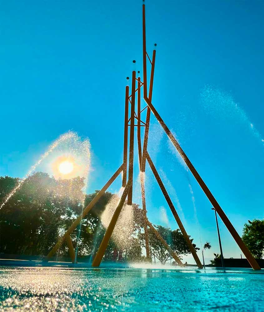 Chafariz com estrutura metálica amarela jorrando água em diferentes direções, iluminado pelo sol em dia claro no Parque da Pavuna, Rio de Janeiro.