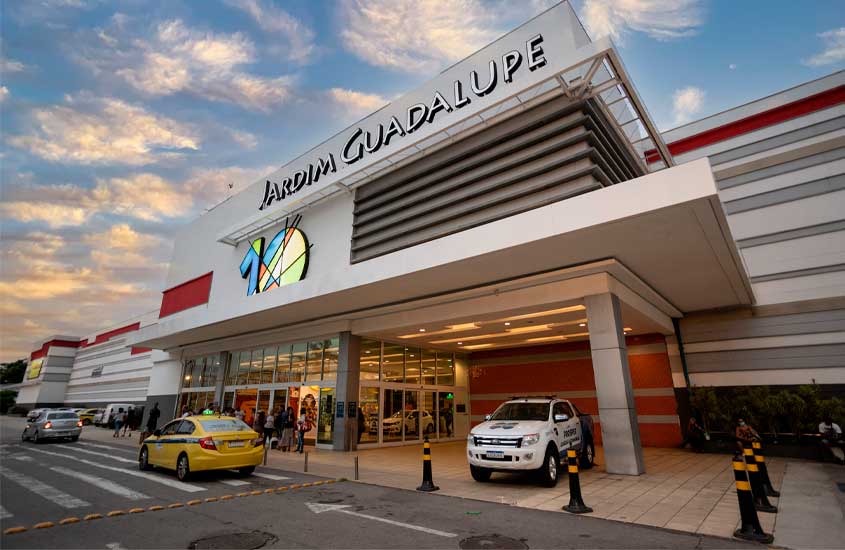 Fachada do Shopping Jardim Guadalupe, no Rio de Janeiro, com entrada principal iluminada ao entardecer, pessoas circulando e carros estacionados em frente