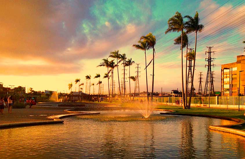 Parque urbano ao pôr do sol, com lago artificial, chafariz e palmeiras altas, sob um céu colorido entre dourado e azul.