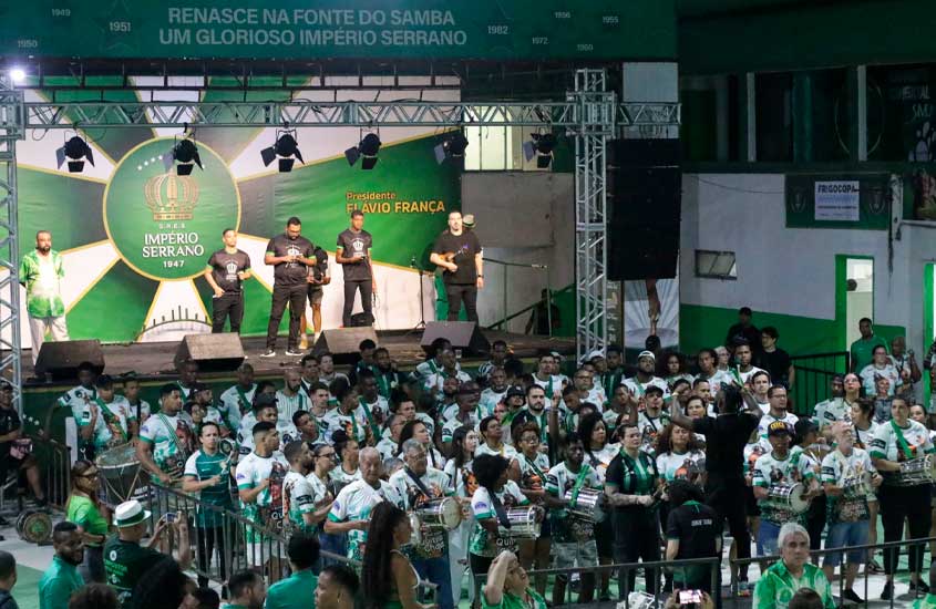 Ensaio da escola de samba Império Serrano em sua quadra, com integrantes da bateria vestidos em verde e branco e músicos no palco sob o estandarte da agremiação.