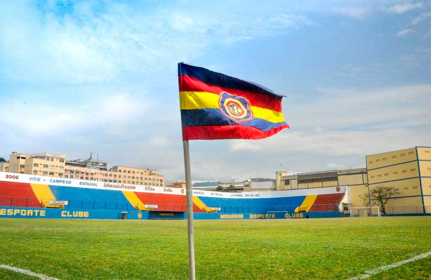 Campo do Estádio Aniceto Moscoso, em Madureira, com bandeira do Madureira Esporte Clube em destaque e arquibancadas coloridas em azul, vermelho e amarelo ao fundo.
