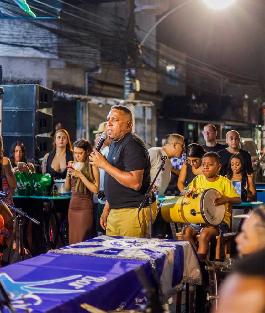 Roda de samba no Pagode da Tia Doca, em Madureira, com cantor ao microfone e menino tocando tamborim, cercados pelo público em clima de festa popular.