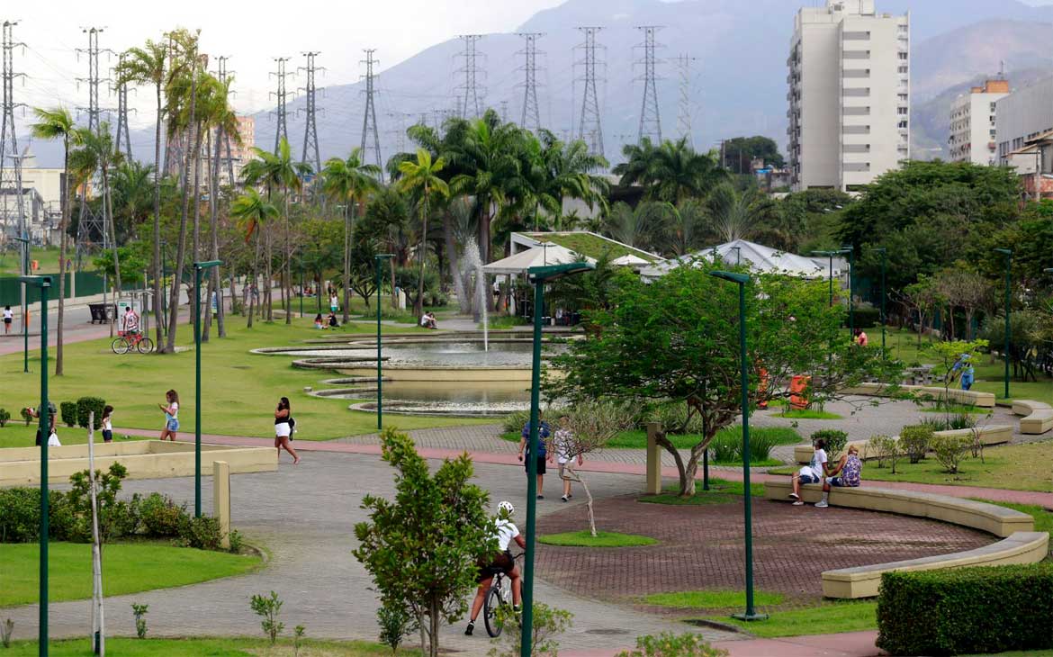 Área verde do Parque Madureira, no Rio de Janeiro, com chafariz, palmeiras, ciclovia e pessoas caminhando e pedalando em meio aos jardins.
