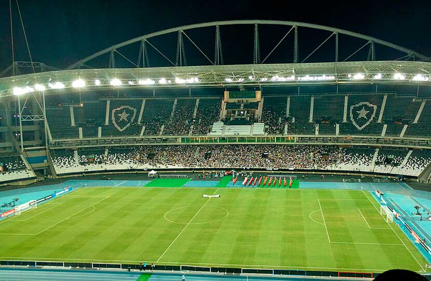 Estádio Nilton Santos, no Rio de Janeiro, conhecido como Engenhão, iluminado à noite durante um jogo de futebol com torcida presente nas arquibancadas.