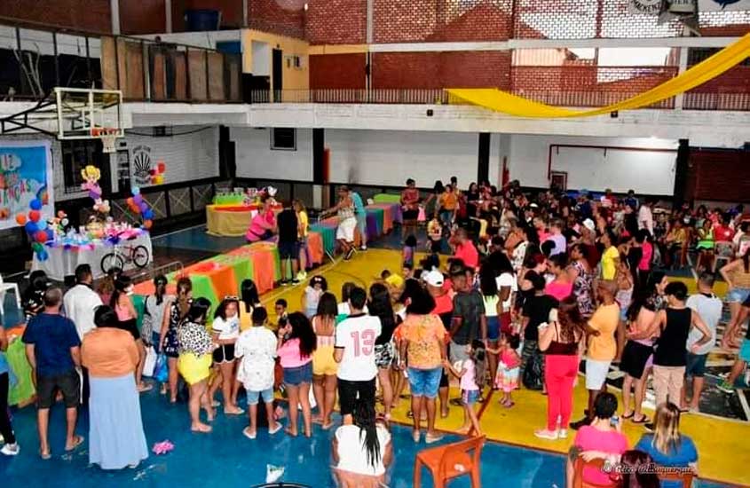Evento comunitário no Clube Mackenzie, no Méier, Rio de Janeiro, com famílias e crianças reunidas em quadra coberta decorada com mesas coloridas.