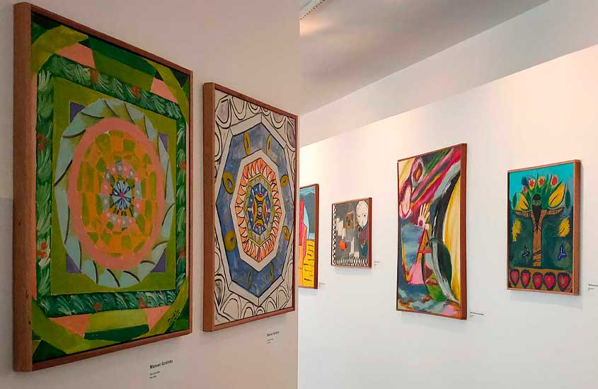 Exposição de arte em museu, com pinturas coloridas em diferentes estilos expostas em paredes brancas iluminadas.