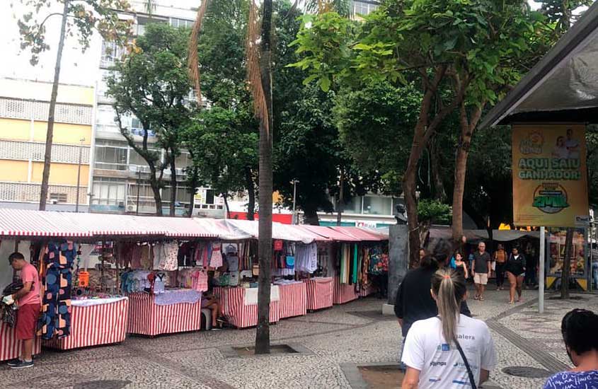 Feira de rua no Méier, com barracas de lona listrada em vermelho e branco vendendo roupas e outros produtos, instalada em uma calçada arborizada com movimento de pedestres.