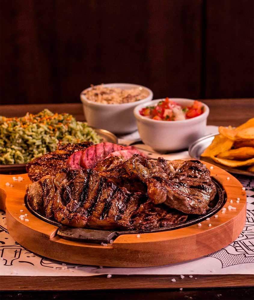 Tábua de carnes assadas com linguiça servida em churrascaria, acompanhada de arroz, farofa, vinagrete e batatas fritas.