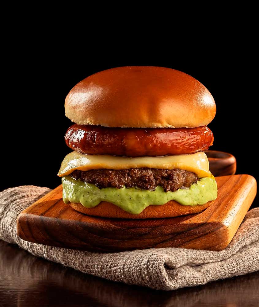 Hambúrguer artesanal servido em pão brioche, com linguiça grelhada, queijo derretido, hambúrguer de carne e molho cremoso verde.