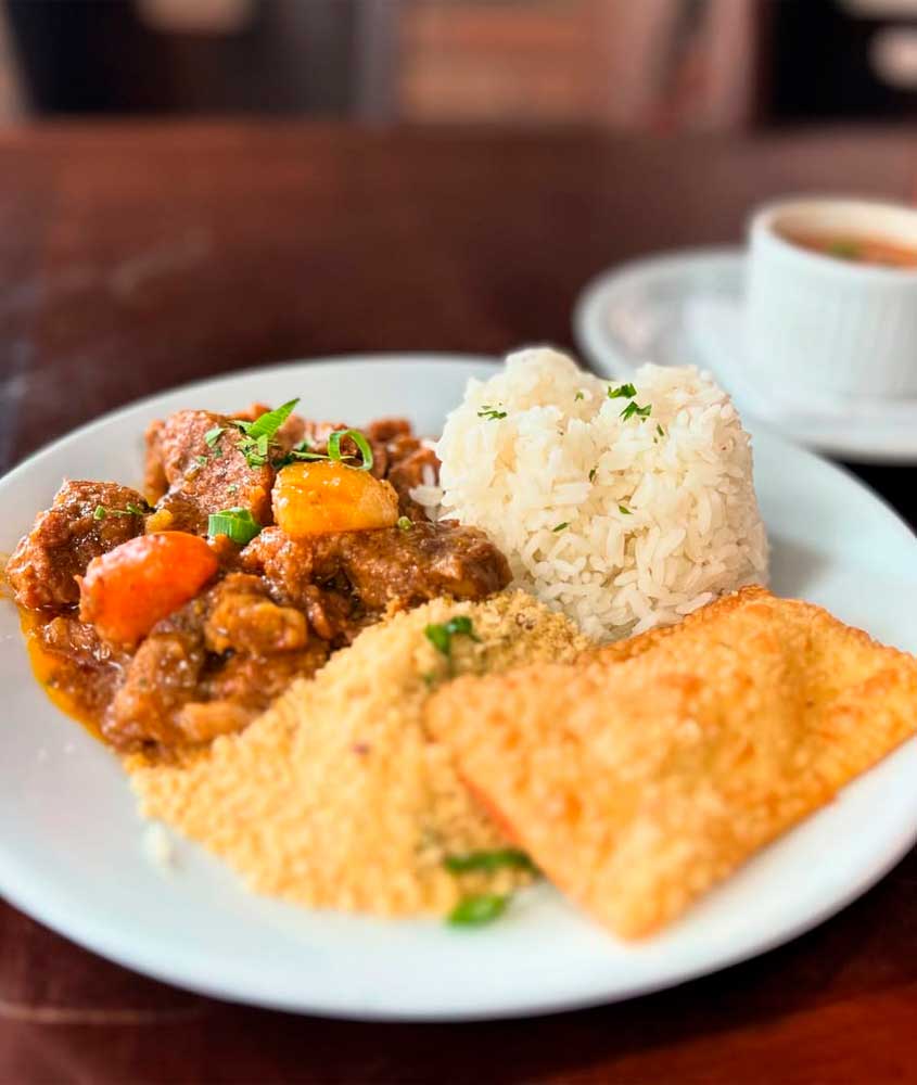 Prato feito com arroz, ensopado de carne com legumes, farofa e pastel frito, acompanhado por uma xícara de caldo.