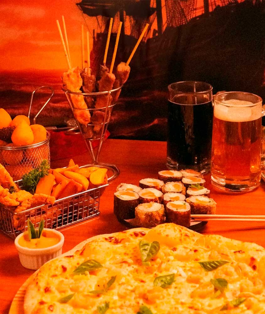 Pizza com queijo e manjericão, acompanhada por espetinhos, coxinhas, batatas fritas, sushi, molho e bebidas (refrigerante e chope) sobre a mesa.