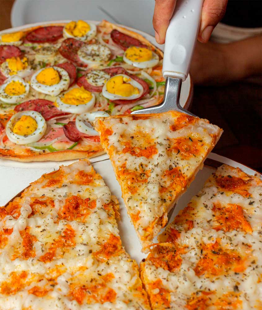Duas pizzas sobre a mesa: uma de queijo com frango desfiado e outra de calabresa com ovo cozido, cebola e pimentão. Uma fatia está sendo servida com espátula.