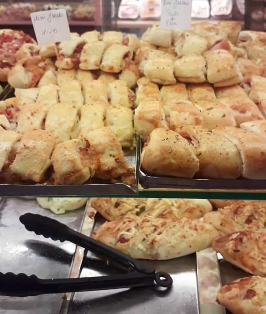 Balcão de padaria com bandejas cheias de enroladinhos salgados assados, cobertos com queijo e temperos. Há plaquinhas indicando preços e tipos de produto, e uma pinça preta à frente para servir os clientes.