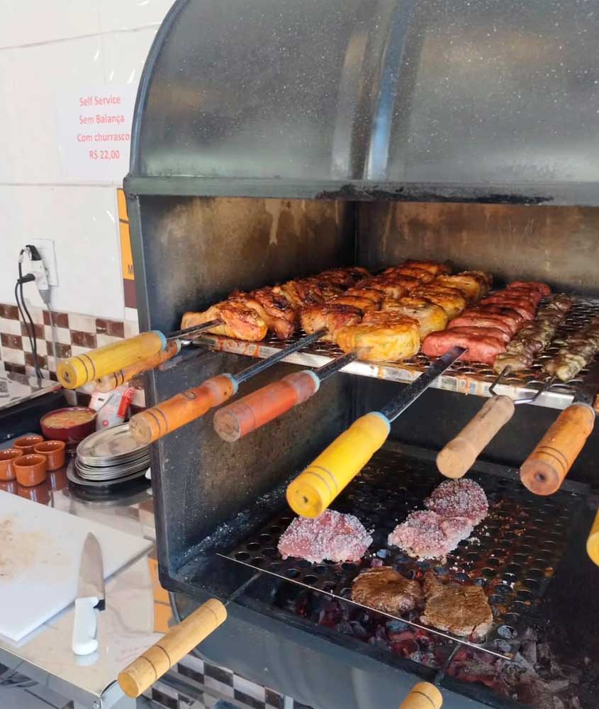 Churrasqueira com espetos de carnes variadas, incluindo frango, linguiça e miúdos, assando sobre brasa. Ao lado, há uma bancada com faca, tábua de corte, pratos e copos, além de uma placa informando o valor do self-service com churrasco.