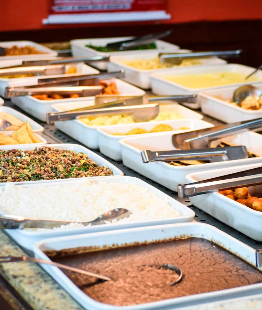 Balcão de self-service com diversas travessas de comida, incluindo arroz, feijão, farofa, legumes, frituras e outros acompanhamentos, organizados em recipientes brancos com colheres e pegadores de metal.