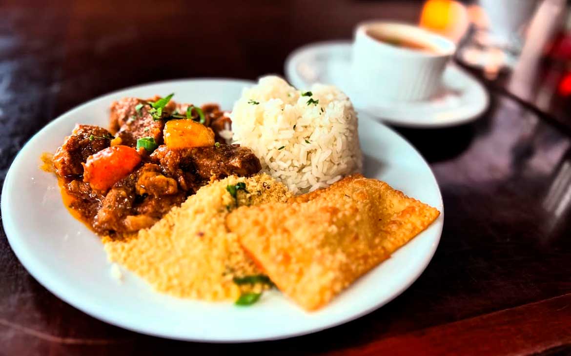Prato de comida brasileira servido em restaurante, com arroz, ensopado de carne com legumes, farofa e pastel.
