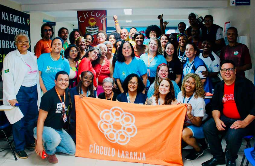 Grupo diverso de pessoas reunidas em espaço interno, sorrindo e segurando uma bandeira laranja com o nome e o símbolo do Círculo Laranja.