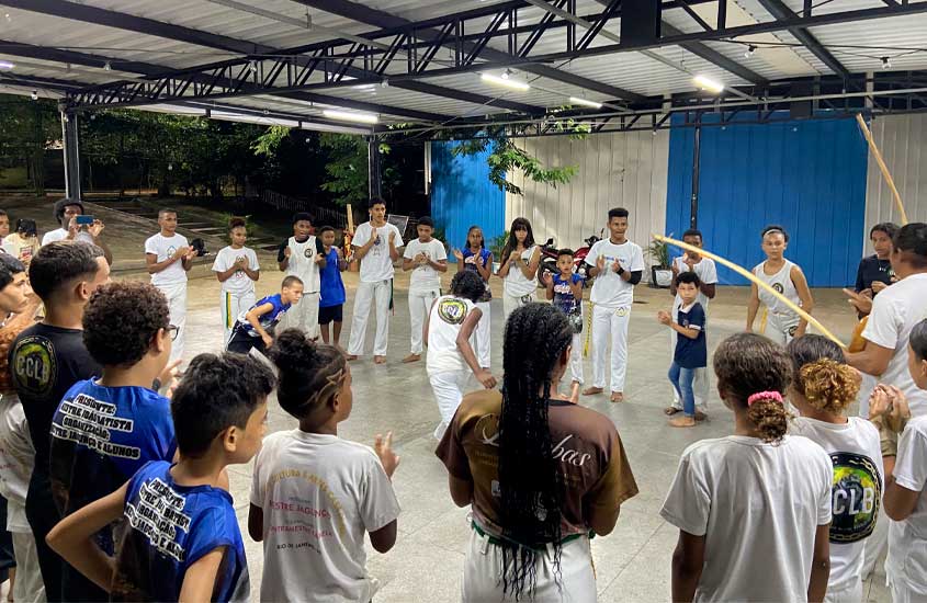 Um grupo de crianças, jovens e adultos forma uma roda de capoeira em um espaço coberto. Algumas pessoas tocam berimbau e batem palmas, enquanto duas crianças jogam capoeira no centro da roda.