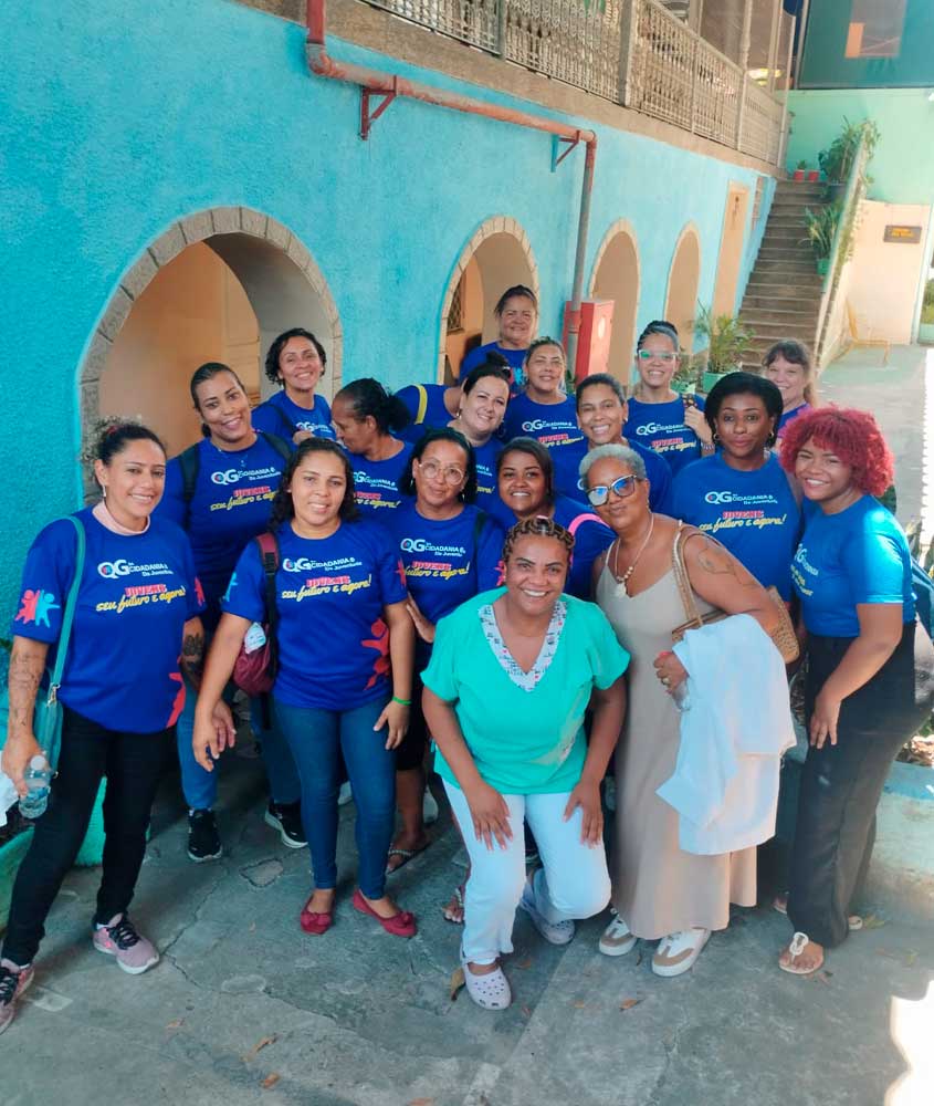 Um grupo de mulheres posa sorridente para a foto em frente a uma parede azul com arcos. A maioria veste camisetas azuis do QG da Cidadania, enquanto duas mulheres ao centro estão com roupas diferentes, uma de verde e branco e outra com vestido bege.