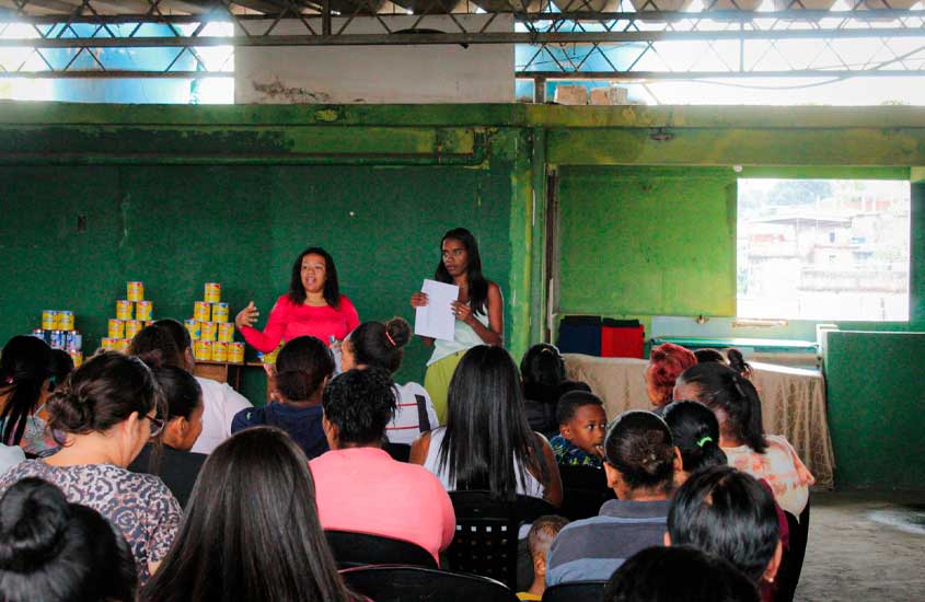 Mulheres participam de uma reunião em um espaço com paredes verdes. Duas palestrantes estão à frente, uma falando e a outra segurando papéis, enquanto o público escuta atentamente. Ao fundo, há pilhas de latas organizadas sobremesas.