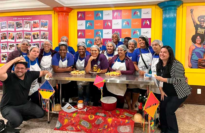 Grupo de pessoas posa sorridente em frente a uma mesa com pratos preparados, algumas vestindo aventais e toucas de cozinha. O ambiente colorido tem bandeirinhas de festa junina como decoração.