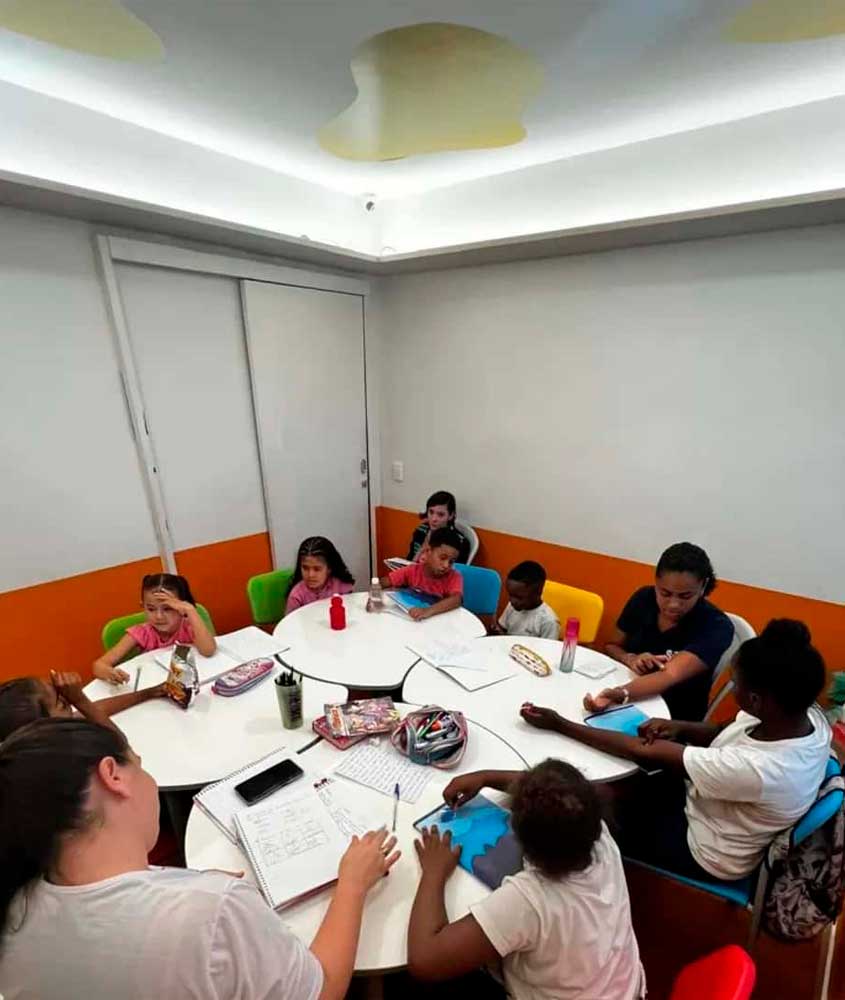 Crianças e uma educadora estão reunidas em uma sala colorida, sentadas ao redor de mesas redondas, fazendo atividades escolares com cadernos, lápis e papéis.