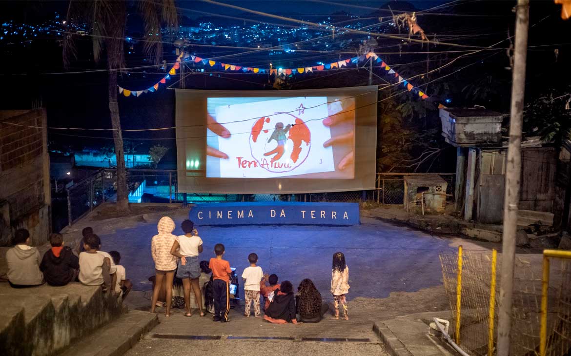 Crianças assistem a uma sessão de cinema ao ar livre diante de uma tela com a escrita “Cinema da Terra”.