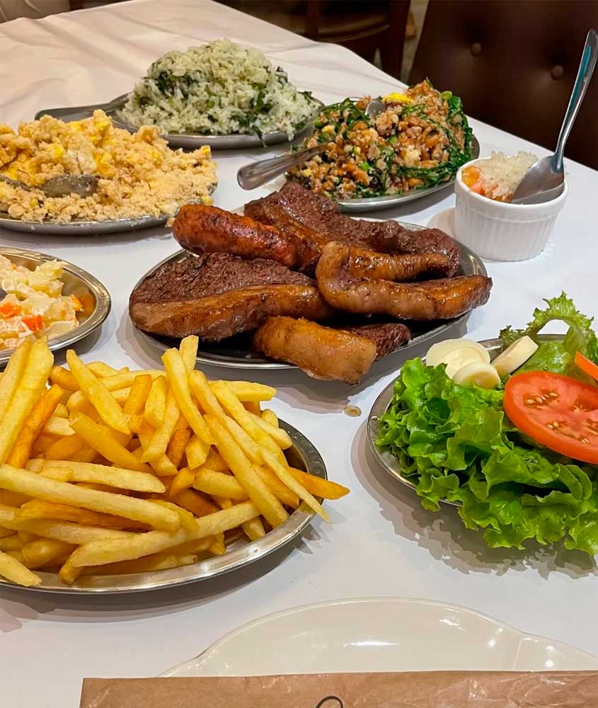 Pratos de churrascaria com picanha assada, linguiça, batata frita, arroz, farofa, saladas e acompanhamentos sobre mesa coberta com toalha branca.