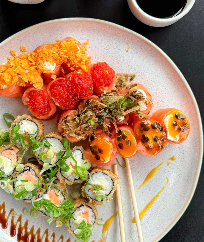 Prato de sushi variado com rolinhos empanados cobertos de cebolinha e sashimis de salmão recheados, decorados com tomate, maracujá e crocantes, acompanhado de molho shoyu.