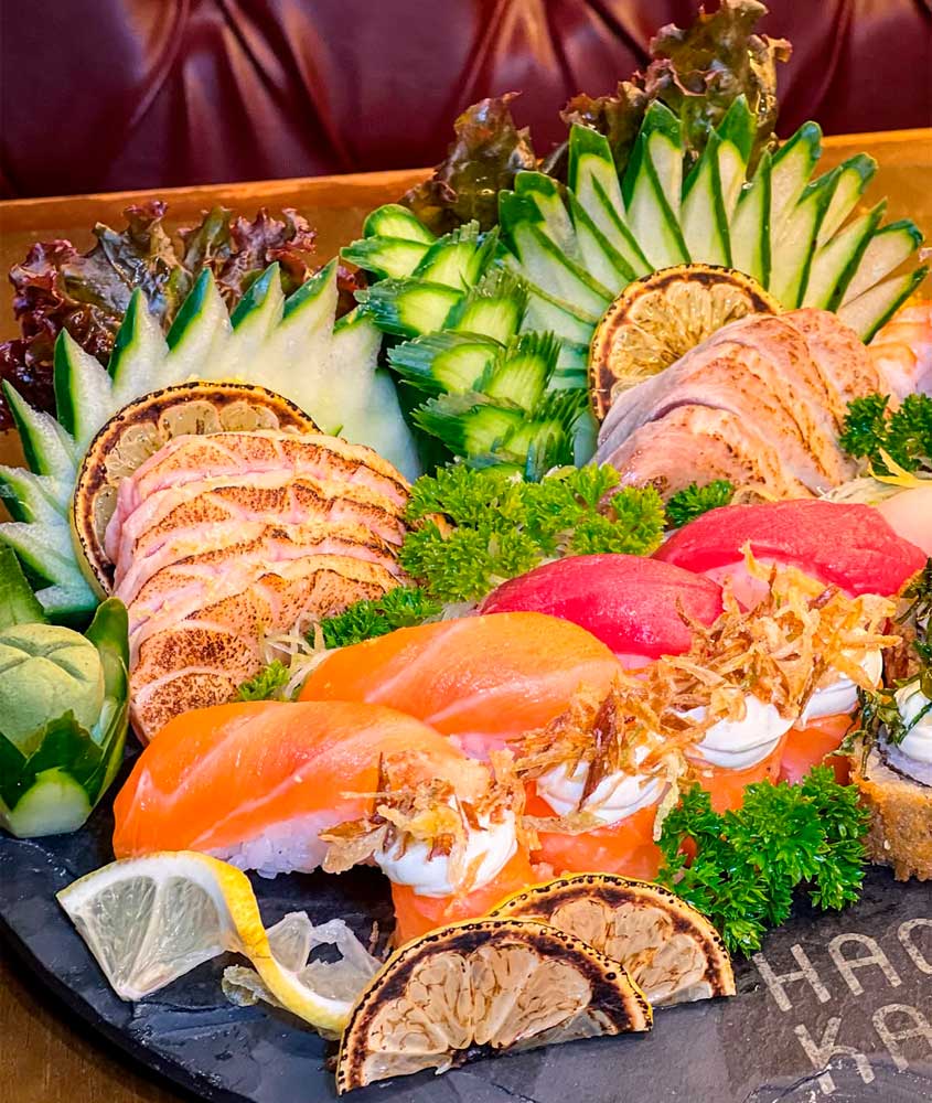 Barca de sushi com sashimis de salmão e atum, niguiris decorados com creme e crocantes, fatias de peixe selado, guarnições de limão queimado e arranjos de pepino e alface.