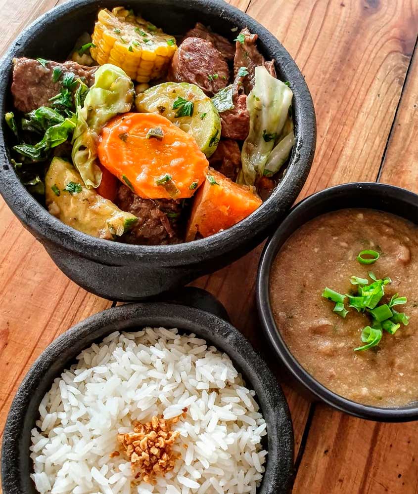 Prato de cozido servido em panela de pedra com carne, linguiça, batata, cenoura, abóbora, milho e repolho, acompanhado de arroz branco com alho frito e feijão em caldo grosso.