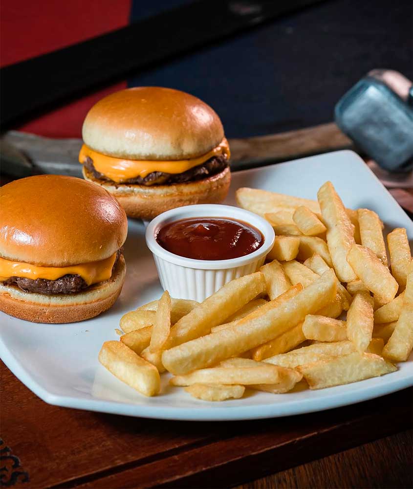 Dois mini-hambúrgueres com queijo cheddar servidos em pão macio, acompanhados de batata frita crocante e molho de ketchup em pote branco.