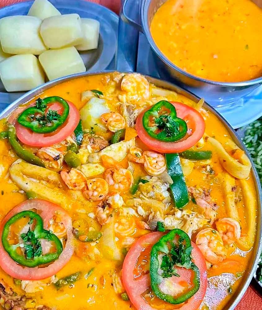 Moqueca de frutos do mar servida em panela com camarões, tomates e pimentões, acompanhada de arroz, mandioca cozida e pirão.