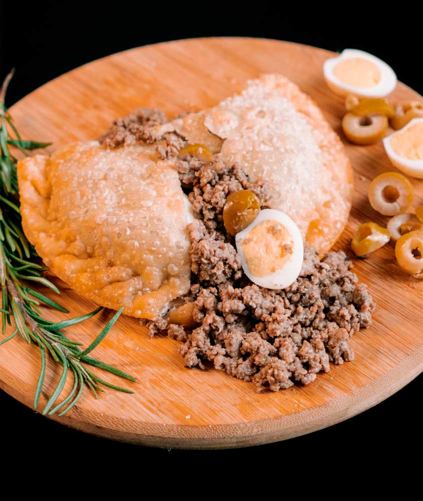 Pastéis recheados acompanhados de carne moída, ovos de codorna e azeitonas verdes sobre tábua de madeira.