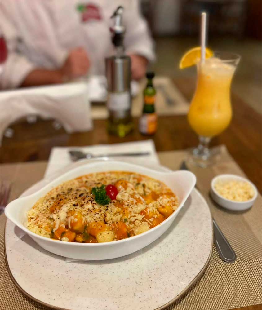 Prato de nhoque ao molho de tomate gratinado com queijo, servido em travessa branca, acompanhado de queijo ralado e bebida cítrica com gelo e fatia de laranja.