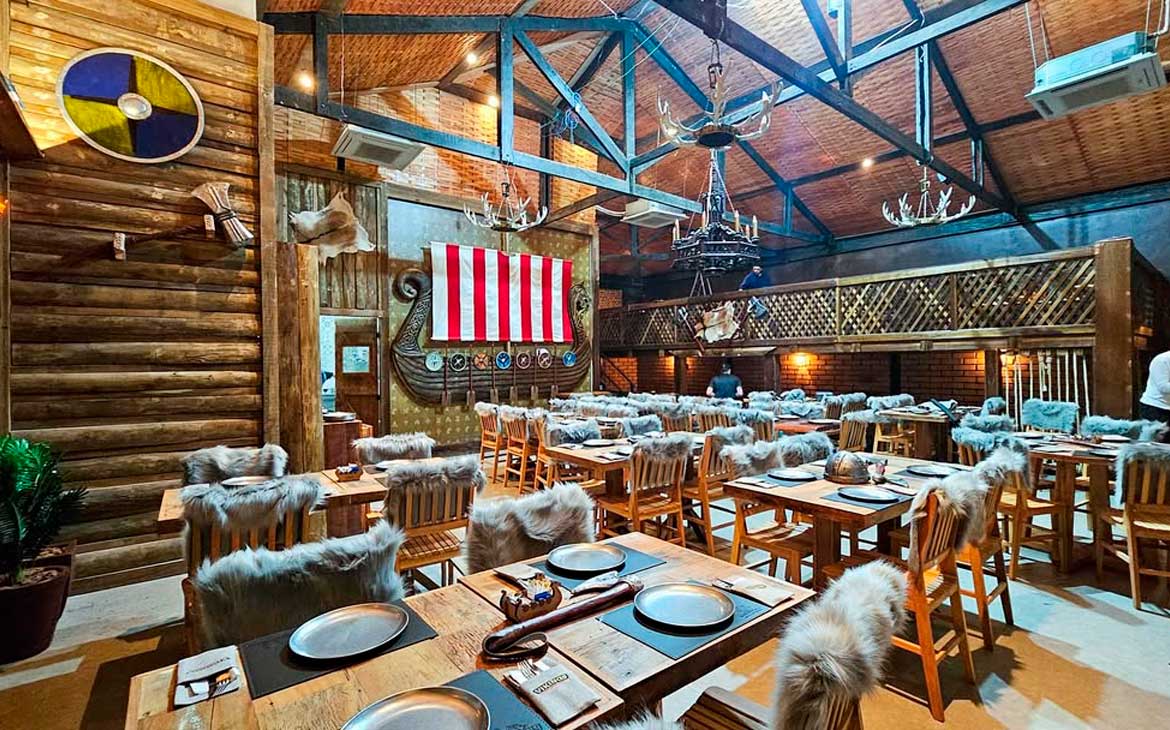 Restaurante temático viking com mesas de madeira, cadeiras cobertas por peles artificiais, decoração de barco com vela listrada vermelha e branca ao fundo e lustres em formato de chifres pendurados no teto.